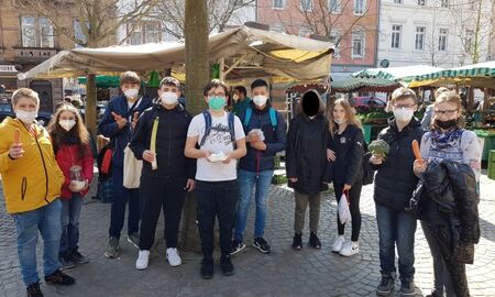 10 Schüler*innen stehen auf dem Markt. Sie haben verschiedene Gemüse in der Hand: Karoote, Brokkoli,... . Im Hintergrund sieht man Marktstände.