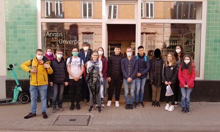 14 Schüler*innen stehen vor dem Laden "Annas Unverpacktes" auf dem Gehweg. Durch die Schaufenster sieht man die Regale mit dem Angebot.