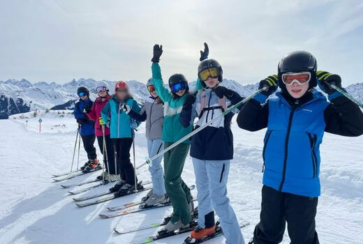 Sieben Jugendliche stehen in Reihe in einer verschneiten Berglandschaft. Sie tragen alle eine Skiausrüstung und lachen in die Kamera. Ein Kind wirft übermütig die Arme in die Höhe.