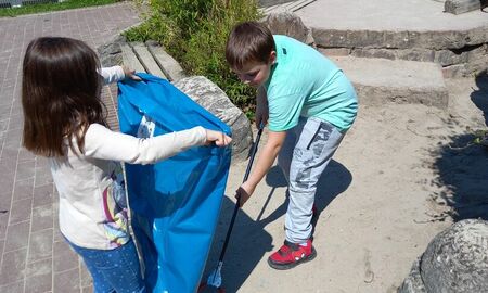Ein Mädchen hält die Mülltüte auf und ein Junge sammelt mit einer Müllzange ein benutztes Taschentuch im Sand neben dem Klettergerüst auf.