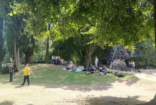 Gruppen von SchülerInnen sitzen auf Picknickdecken oder stehen auf einer Wiese mit hohen Bäumen. Im Hintergrund sieht man das Kletternetz.