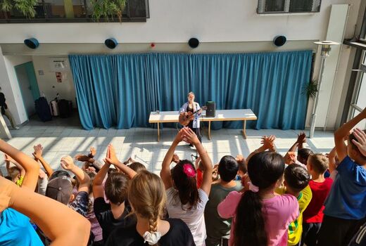 Eine Lehrerin steht mit Gitarre auf der Bühne. Im Vordergrund stehen die Grundschüler, den Rücken zum Betrachter gewandt und klatschen in die Hände.