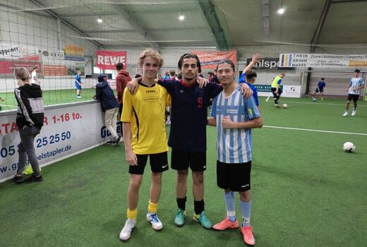 Drei Junge Männer in Fußballoutfit in der Fußballhalle. Sie schauen in die Kamera. Im Hintergrund trainieren SchülerInnen.