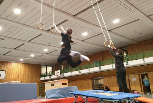 Eine Jugendliche greift im Sprung nach den Ringen. Im Hintergrund schwingt ein Junge an den Ringen.