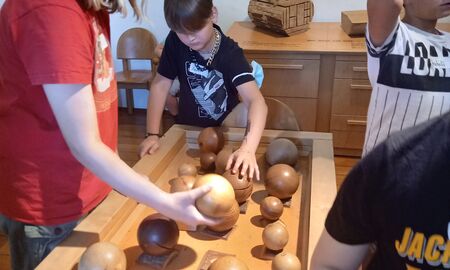 Eine Viertklässlerin und drei Viertklässler testen an einem Ausstellungstisch das Gewicht unterschiedlicher Holzarten mithilfe von Holzkugel aus.