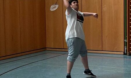 Ein Spieler schaut in die Luft und macht eine Schlagbewegung mit dem Badminton Schläger. Im Hintergrund ist die braune Wand der Turnhalle zu sehen.