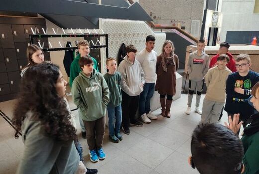 Zwölf Schüler und eine Lehrerin stehen im Rund, eine Person ganz rechts am Bildrand erklärt etwas.