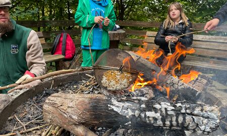 Ein Waldpädagoge hält ein geschlossenes Sieb mit Popcorn übers Lagerfeuer und Kinder schauen ihm dabei zu.