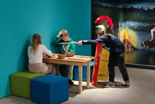 Zwei SchülerInnen sitzen an einem Tisch und spielen ein Spiel. Ein Schüler, verkleidet als Soldat mit Helm, Schwert und Schild greift sie mit dem Gladius an.