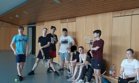 Schülerinnen und Schüler sitzen und stehen beisammen. Sie befinden sich in der Turnhalle. Manche haben einen Badminton Schläger in der Hand