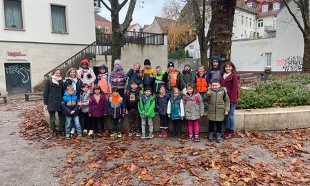 Zwanzig Schülerinnen und Schüler aus zwei ersten Klassen posieren mit zwei Lehrerinnen und einer Erzieherin vor einem Baum in der Stadt für ein Gruppenfoto.