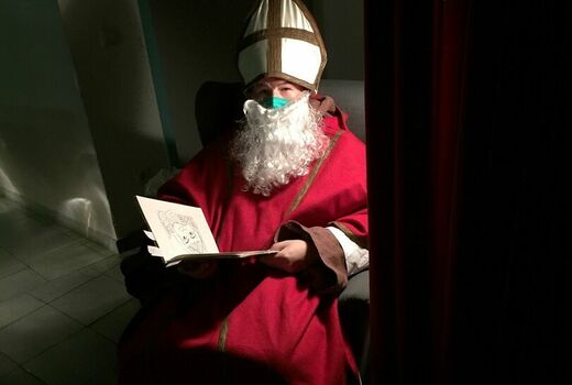 Der Nikolaus sitzt auf einem Sessel. Er hält ein aufgeschlagenes Buch in der Hand. Es ist dunkel und nur der Nikolaus ist beleuchtet.
