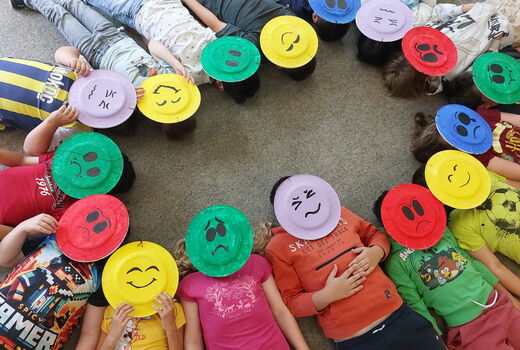 Sechzehn Kinder liegen im Kreis und tragen bunte Masken aus Papptellern, die verschiedene Gefühle in Form von Smileys darstellen.