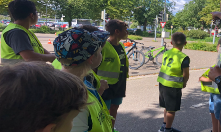 Sieben Kinder in Warnwesten stehen an einer Übungsstraße und hören einer Erklärung aufmerksam zu.
